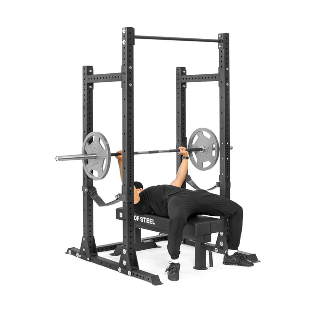 A person lies on a bench press bench, gripping a barbell with weights, inside the Bells of Steel Hydra Half Rack - Prebuilt (3" x 3", ⅝" Holes, 1000-lb capacity), against a plain white background.