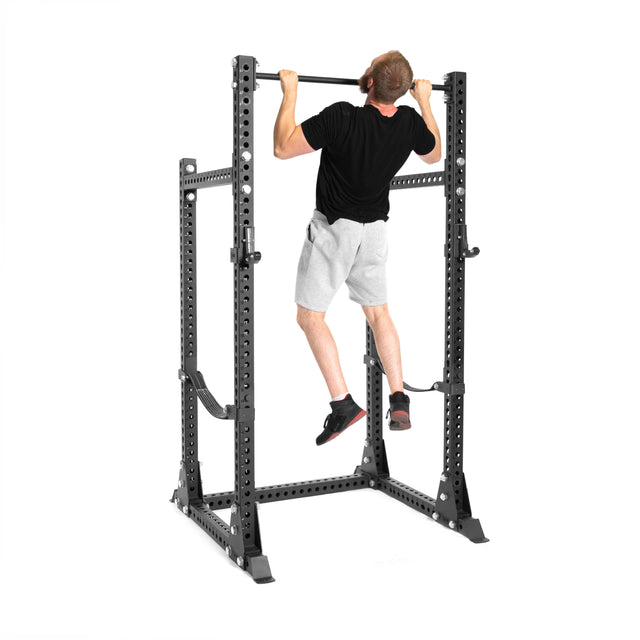 A man in a black t-shirt and gray shorts does a pull-up on the Bells of Steel Manticore Half Rack - Prebuilt (3" x 3", 1" holes), the perfect addition to any home gym, set against a white background.