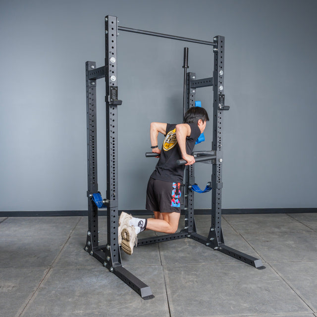 A person in athletic attire does a dip on parallel bars mounted to the Bells of Steel Hydra Half Rack - Prebuilt (3" x 3", ⅝" Holes) in a gym with gray walls and a tiled floor.