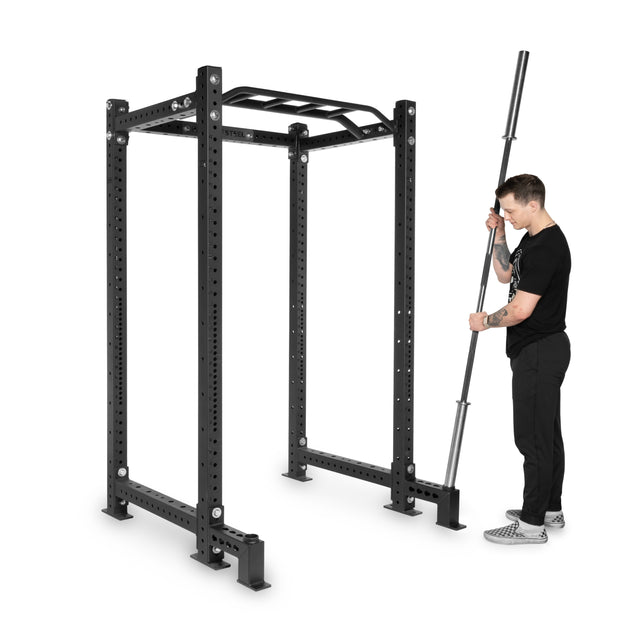A man in black clothes holds a barbell next to a Bells of Steel power rack with Front Foot Stabilizers and a pull-up bar attachment on top.