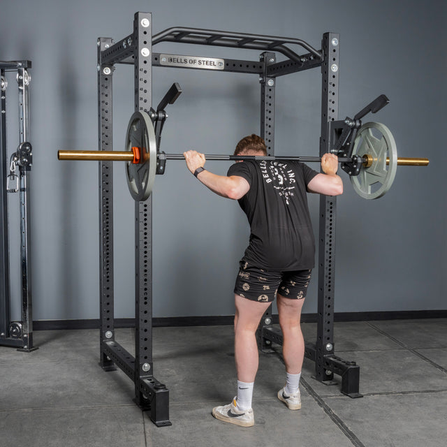 Wearing a black shirt and shorts, a person readies for a barbell squat using the Bells of Steel Monolift Rack Attachment, with weights loaded on each end, inside a gym with gray flooring.