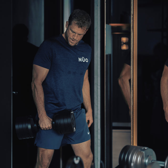 A man in a blue t-shirt and shorts lifts a Bells of Steel NÜOBELL-S Adjustable Dumbbell in the gym, standing near a mirror with a focused expression.
