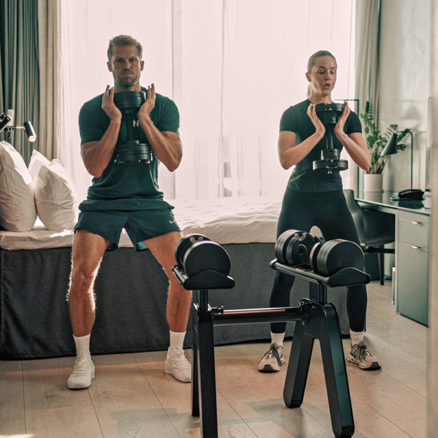 Two people in workout gear perform squats in a home gym, each holding NÜOBELL-S Adjustable Dumbbells by Bells of Steel. A dumbbell rack stands in the foreground as sunlight streams through a large window behind them.