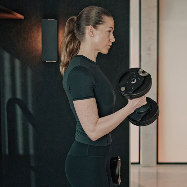A woman with a ponytail in black athletic wear does a bicep curl using Bells of Steel NÜOBELL-S Adjustable Dumbbells in a sleek, modern home gym.