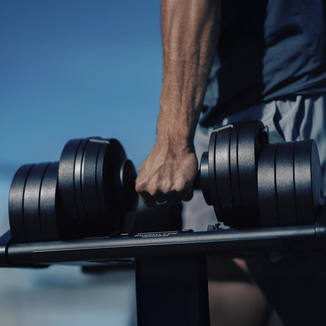 A muscular arm grasps a Bells of Steel NÜOBELL-S Adjustable Dumbbell on a rack, with a blue sky in the background, highlighting the product’s outdoor strength training versatility and precision engineering.