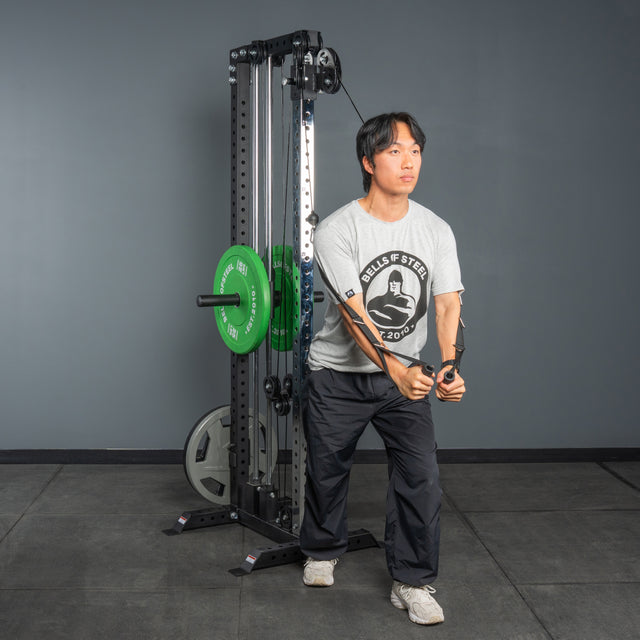 A man in a gray T-shirt and black pants performs a cable chest fly using the Bells of Steel Cable Tower, standing in front of it with green and black weight plates at the gym.
