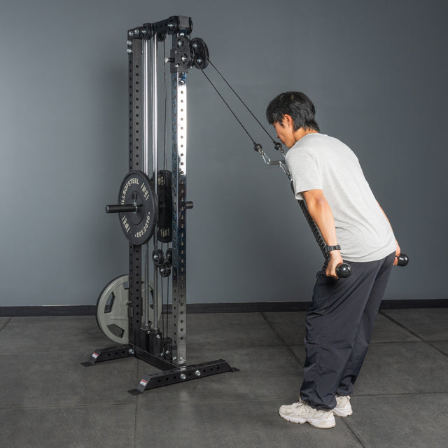 A person in a gray t-shirt and black pants uses the Bells of Steel Cable Tower’s weight stack to perform a triceps pushdown in a gym with gray walls and flooring.