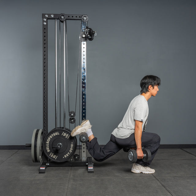 A person does a Bulgarian split squat with one foot on the Bells of Steel Cable Tower, holding dumbbells in each hand, set against a gray gym background.