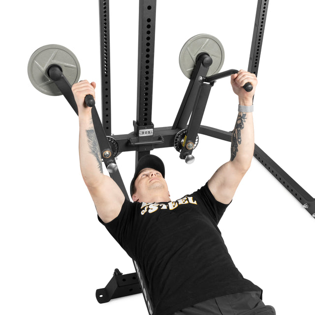 A person in a black cap and t-shirt uses the Bells of Steel Shoulder Boulder/Chest Fly Attachment, gripping its handles and lying on a bench mid-workout.