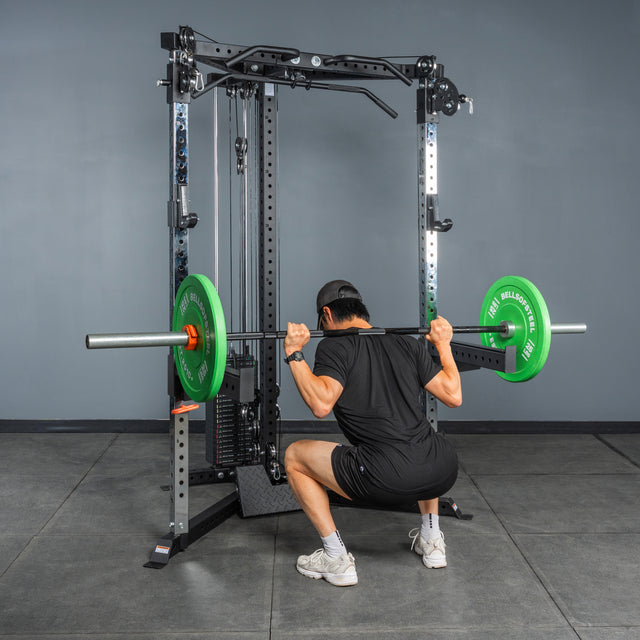 A person uses the Bells of Steel All-in-One Trainer, wearing a black shirt, shorts, and cap, to perform a barbell squat with green plates inside a squat stand in the gym.