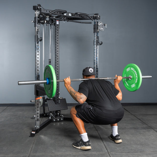 Wearing black and a cap, a person performs a barbell squat with green plates on the Bells of Steel All-in-One Trainer squat rack in a gym with gray concrete floors and walls.