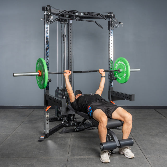 Wearing athletic gear, a person bench presses with a barbell and green plates on a bench inside a gym, using the Bells of Steel All-in-One Trainer power rack and squat stand on a gray tiled floor.