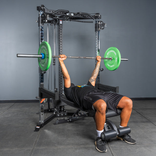 A person bench presses a barbell with green weight plates inside the Bells of Steel All-in-One Trainer in a gym with gray walls and flooring.