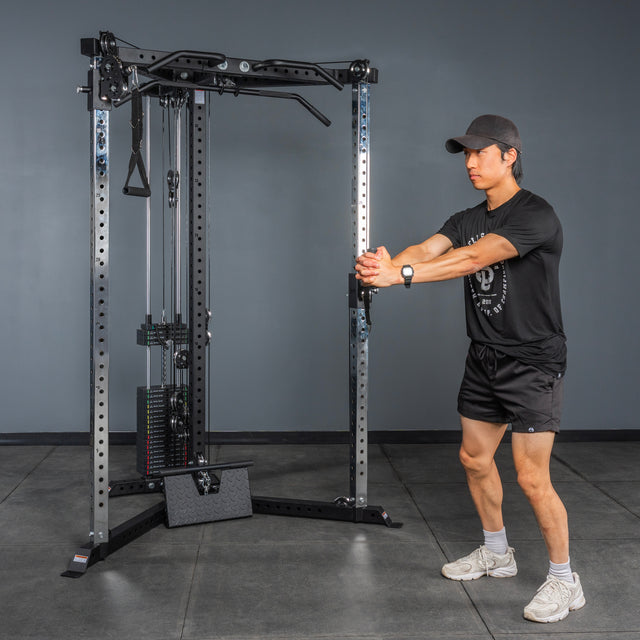 A person in athletic wear prepares to perform a cable exercise with the Bells of Steel All-in-One Trainer, holding the handle with both hands extended forward in a gym or home gym setting.