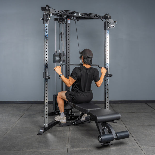 Wearing black workout clothes, a person uses the Bells of Steel All-in-One Trainer to perform a lat pulldown while seated on an adjustable bench in a gym with gray walls and flooring.