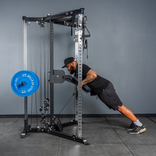 A man in black workout gear performs an incline push-up using the Bells of Steel All-in-One Trainer with a blue 45 lb plate in a gym with gray walls and floors.