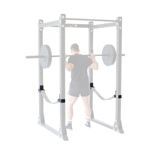 Wearing athletic gear, a person performs a barbell squat inside a rack equipped with Bells of Steel Safety Strap Connectors. The rack’s uprights are subtly faded to draw focus to the lifter.