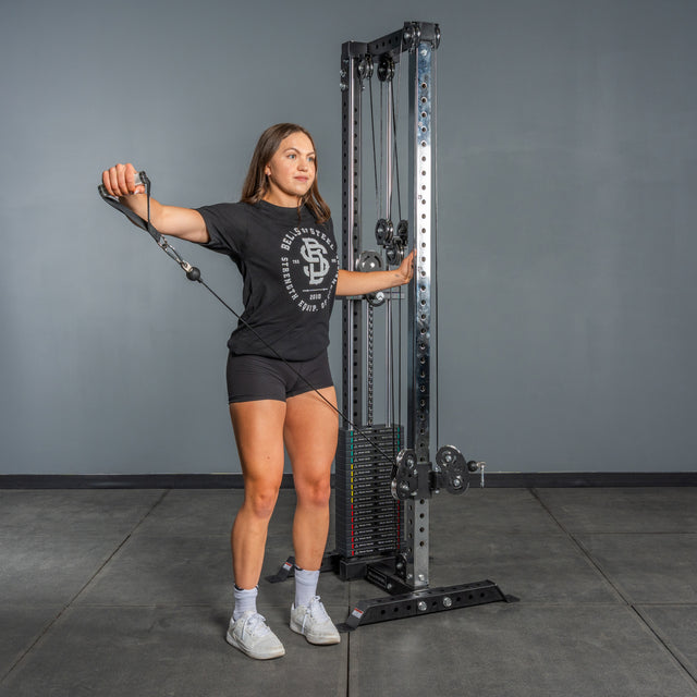 A woman in a black t-shirt, shorts, and sneakers stands in a gym by the Bells of Steel Cable Tower, gripping its handle attached to the weight stack as she prepares for her cable machine workout.