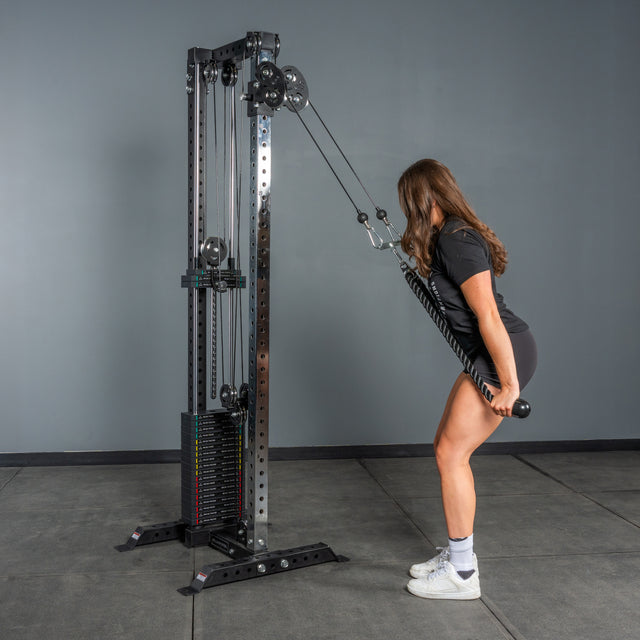 A woman in athletic wear does a triceps pushdown with a rope attachment and weight stack on the Bells of Steel Cable Tower, set in a gym with gray walls and floors.