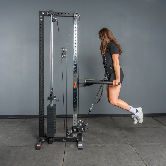 A woman in athletic wear performs a weighted dip exercise on the Bells of Steel Cable Tower in a gym with gray walls and floor tiles.