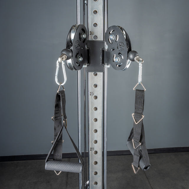 A close-up of Bells of Steel Cable Tower’s adjustable cable pulley attachments with handles. Numbered holes on the vertical post allow precise height changes for various exercises. Set against a gray wall and floor.