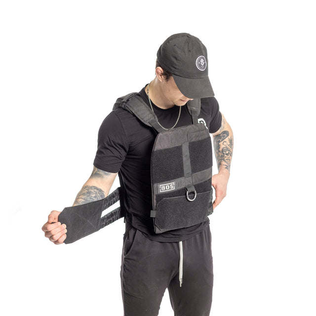 A man in black attire adjusts the strap of the Bells of Steel Weighted Vest and Plates, featuring an adjustable fit, with the brandâs logo displayed on front against a plain white background.
