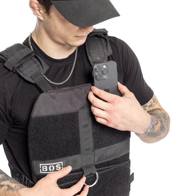 A tattooed person in a black t-shirt, chain, and black cap slips a phone into the front pocket of the Bells of Steel Weighted Vest and Plates, showing off this workout accessoryâs adjustable fit.
