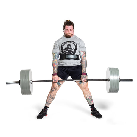 A muscular man with tattoos and a beard lifts a heavy barbell during a deadlift. He wears a "Built of Steel" t-shirt, shorts, a lifting belt, and wrist wraps, standing on a white background.