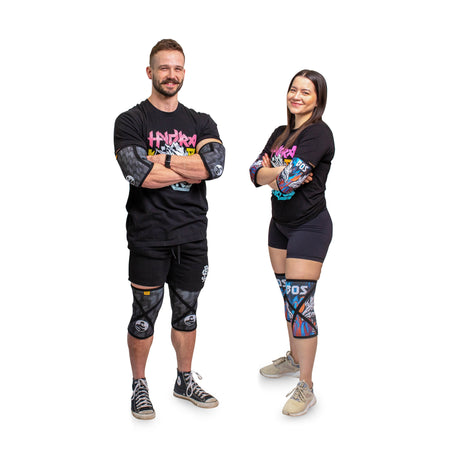 Two people stand side by side, smiling with arms crossed. Both wear black athletic outfits, knee sleeves, and colorful arm sleeves. The background is plain white.