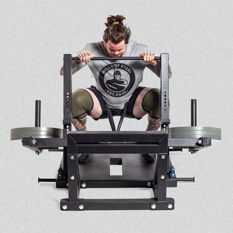A person with a bun and tattoos wearing a "Bells of Steel" T-shirt and knee sleeves is lifting weights on a squat machine with weights loaded on both sides. The background is plain gray.