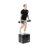 A person performs step-ups with dumbbells on a black Bells of Steel Foam Soft Plyo Box (16 x 18 x 20), wearing a black T-shirt, gray shorts, white socks, and sneakers against a plain white background.