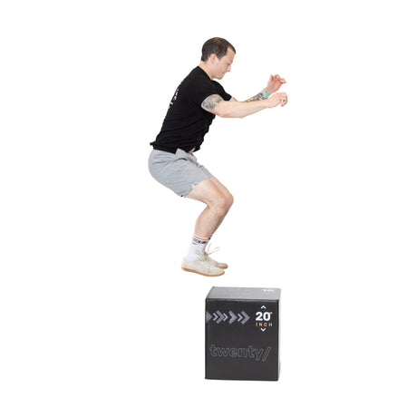 A man in a black shirt and gray shorts is mid-air, executing a box jump onto a Bells of Steel Foam Soft Plyo Box (16 x 18 x 20) with bent arms and tucked knees against a plain white background, showcasing this versatile gym equipment.
