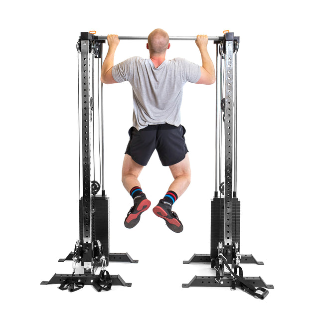 A man in a gray t-shirt, black shorts, and colorful socks performs a pull-up on the Bells of Steel Cable Tower Squat Stands, photographed from behind against a white background.