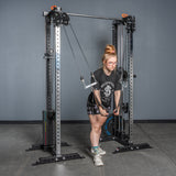 A person uses the Bells of Steel Cable Tower Squat Stands in a gym, performing a cable triceps extension. They wear glasses, a black T-shirt, shorts, and sneakers while standing on a concrete floor in front of a gray wall.