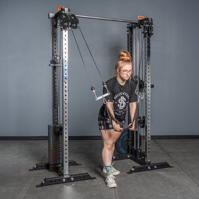 A person uses the Bells of Steel Cable Tower Squat Stands in a gym, performing a cable triceps extension. They wear glasses, a black T-shirt, shorts, and sneakers while standing on a concrete floor in front of a gray wall.