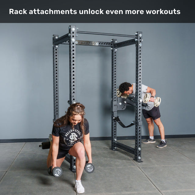 Two people work out with dumbbells using the Bells of Steel Manticore Four Post Power Rack Builder (3" x 3", 1" Holes) in a gym. One person lunges in front while the other performs bent-over lifts at the rack. Text above reads, "Rack attachments unlock even more workouts.