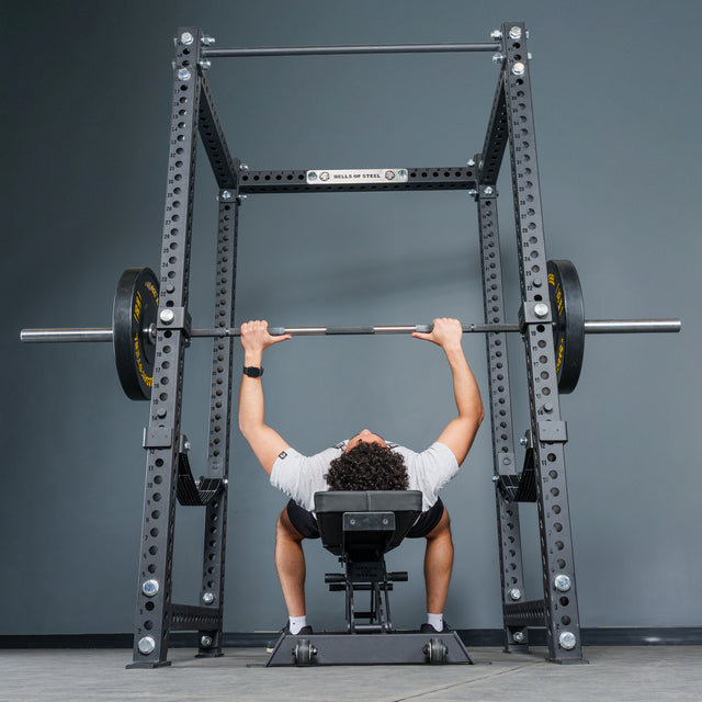 A person lies on a bench inside the Bells of Steel Manticore Four Post Power Rack Builder (3" x 3", 1" Holes), gripping a barbell loaded with weight plates above their chest as if preparing to perform a bench press.