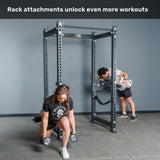 A woman performs lunges with dumbbells in front of the Bells of Steel Manticore Four Post Power Rack - Prebuilt (3" x 3", 1" Holes), while a man uses a preacher curl attachment on the rack. Text above reads, "Rack attachments unlock even more workouts.