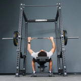 A person lies on a bench press, gripping a barbell loaded with weights, preparing to lift inside the Bells of Steel Manticore Four Post Power Rack – Prebuilt (3" x 3", 1" Holes) positioned against a gray wall.