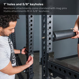 A man adjusts a metal pin on the Bells of Steel Manticore Four Post Power Rack - Prebuilt (3" x 3", 1" holes), while another person holds a cylindrical pad attachment nearby—making it the perfect addition to any well-equipped home gym.