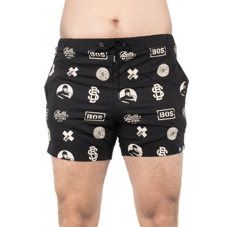 A person stands against a white background wearing black Bells of Steel Gym Shorts with white logos, "BOS," brand text, and graphic designs made from high-performance materials.
