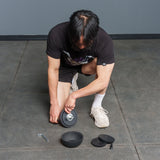 A person kneels on a tiled floor assembling the Bells of Steel 6-12KG Adjustable Kettlebell, with plates, a wrench, and a screw-on collar laid out nearby—a detailed setup reminiscent of demonstrations by Mark Wildman when showcasing adjustable kettlebell equipment.