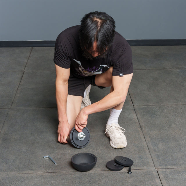 A person kneels on a tiled floor assembling the Bells of Steel 6-12KG Adjustable Kettlebell, with plates, a wrench, and a screw-on collar laid out nearby—a detailed setup reminiscent of demonstrations by Mark Wildman when showcasing adjustable kettlebell equipment.
