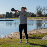 A woman in athletic wear performs a kettlebell swing exercise outdoors by a lake on a sunny day, using the Bells of Steel 6-12KG Adjustable Kettlebell, with trees and houses visible in the background.