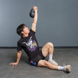 A person in athletic attire performs a Turkish get-up exercise, holding the Bells of Steel 6-12KG Adjustable Kettlebell overhead with one arm while supporting their body with the other arm, sitting on a gym floor and demonstrating a technique popularized by experts like Mark Wildman.