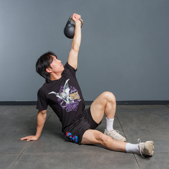 A person in athletic attire performs a Turkish get-up exercise, holding the Bells of Steel 6-12KG Adjustable Kettlebell overhead with one arm while supporting their body with the other arm, sitting on a gym floor and demonstrating a technique popularized by experts like Mark Wildman.