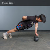 Wearing workout clothes, a person performs a one-arm plank on the Bells of Steel 6-12KG Adjustable Kettlebell, with their other hand behind their back, on a gray-tiled gym floor. Text at top reads “Stable base.”.