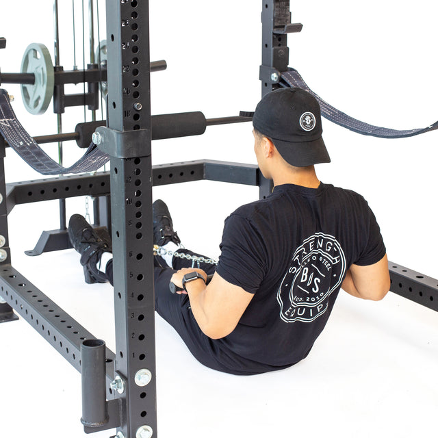 Wearing black athletic wear and a cap, a person sits on the floor, preparing to use the Bells of Steel Lat Pulldown & Low Row Rack Attachment - Hydra & Manticore in a gym with weightlifting equipment and Olympic weight plates.