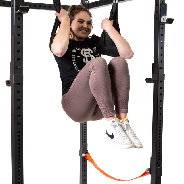A woman smiles while doing a core workout hanging knee raise on a pull-up bar using Bells of Steel Hanging Ab Straps (Pair). She wears a black T-shirt, mauve leggings, and white sneakers.