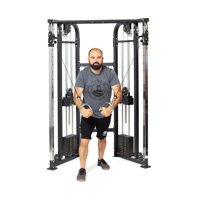 A man uses the Bells of Steel Functional Trainer - Weight Stack, gripping handles with both hands. He wears a gray t-shirt, black shorts, and athletic shoes, standing in a staggered stance against a white background.